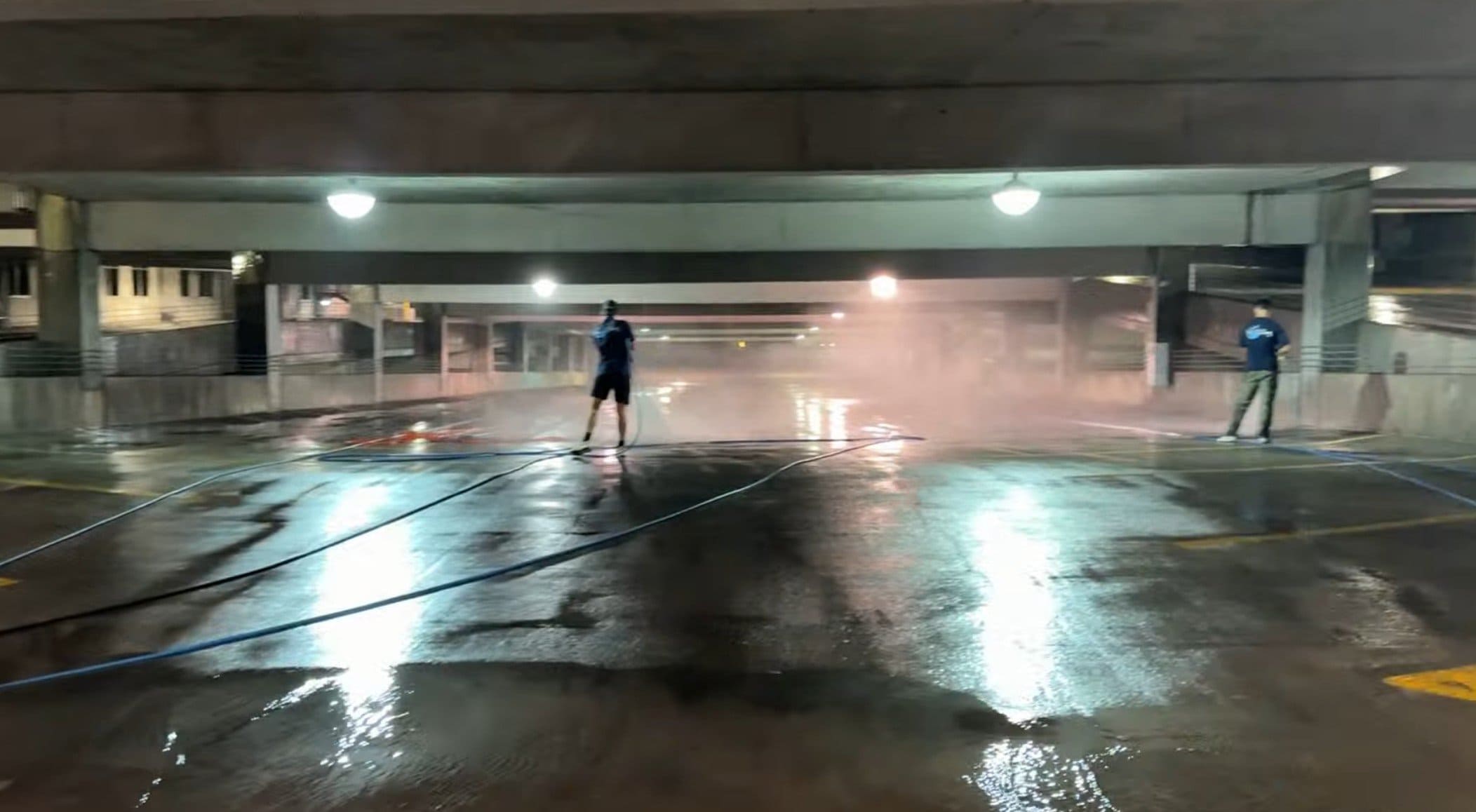 Two KC Splash technicians pressure washing K-State University parking garage at night with heavy steam filling the open level